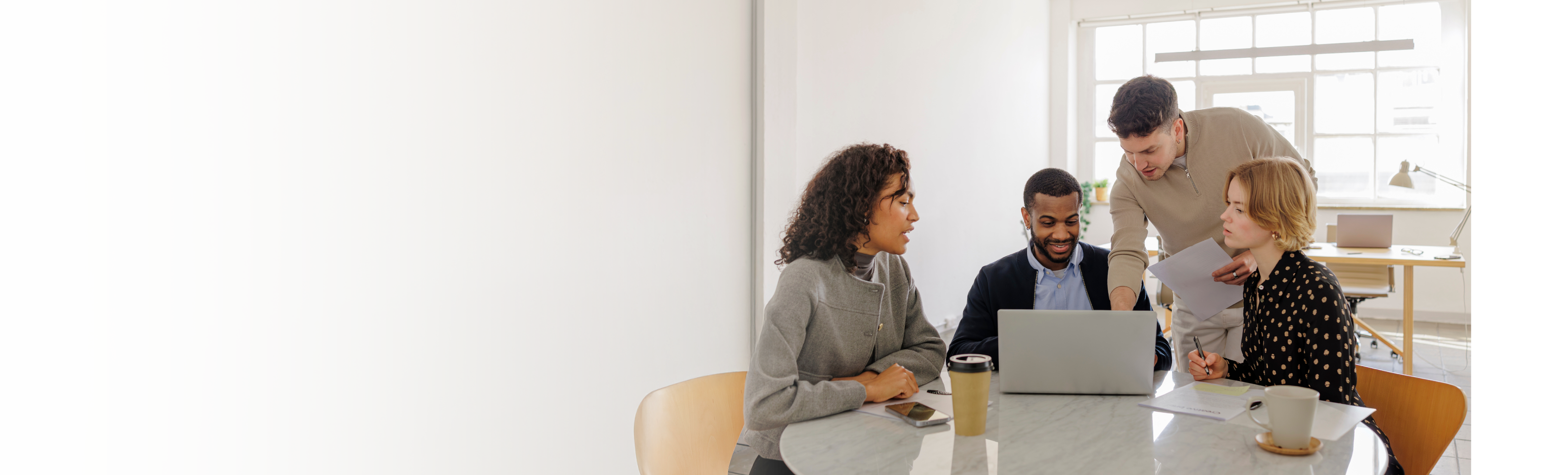 Four coworkers in an office sitting and standing around a table with laptops talking to each other