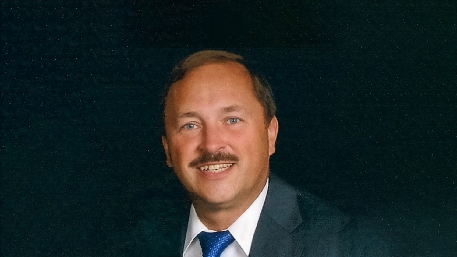 A man dressed in a formal dark suit paired with a white shirt and a vibrant blue tie. The background is a solid dark color, creating a professional and polished appearance. The image conveys a corporate or business-oriented setting.