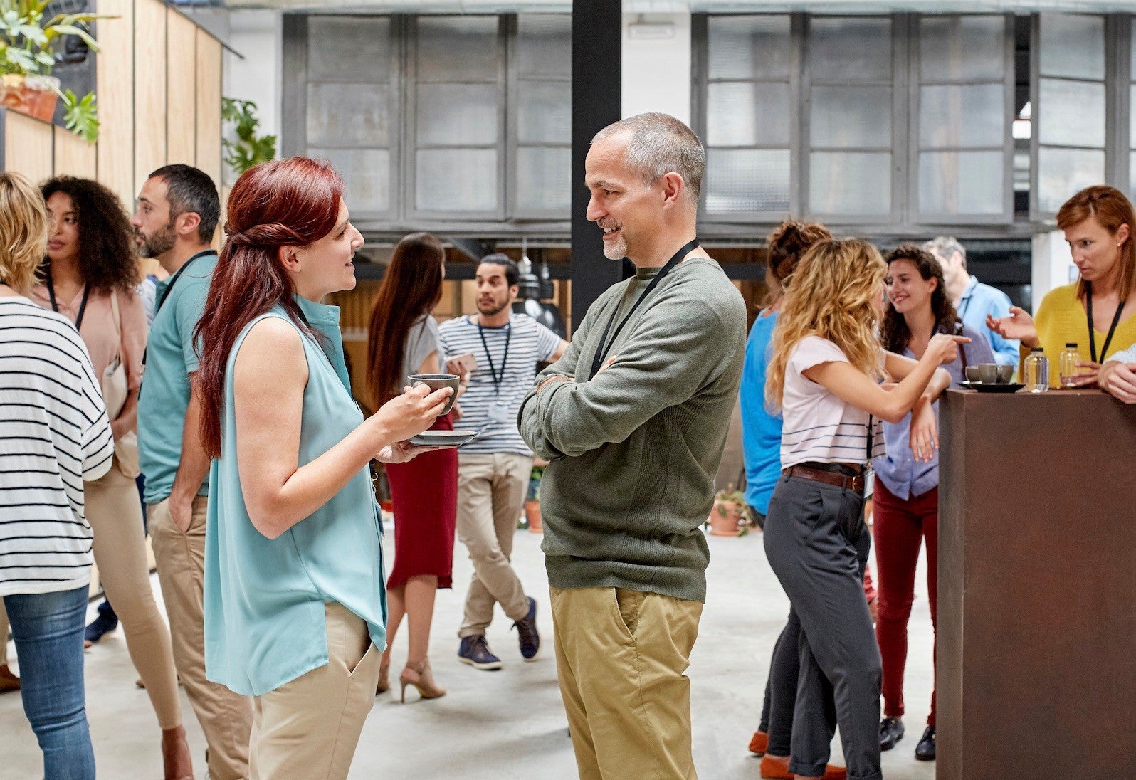A group of people engaging in conversations at a casual networking event held in a modern indoor venue. The setting features natural light, industrial-style windows, and a mix of casual and business-casual attire. Attendees are seen socializing in small groups, creating a lively and interactive atmosphere.