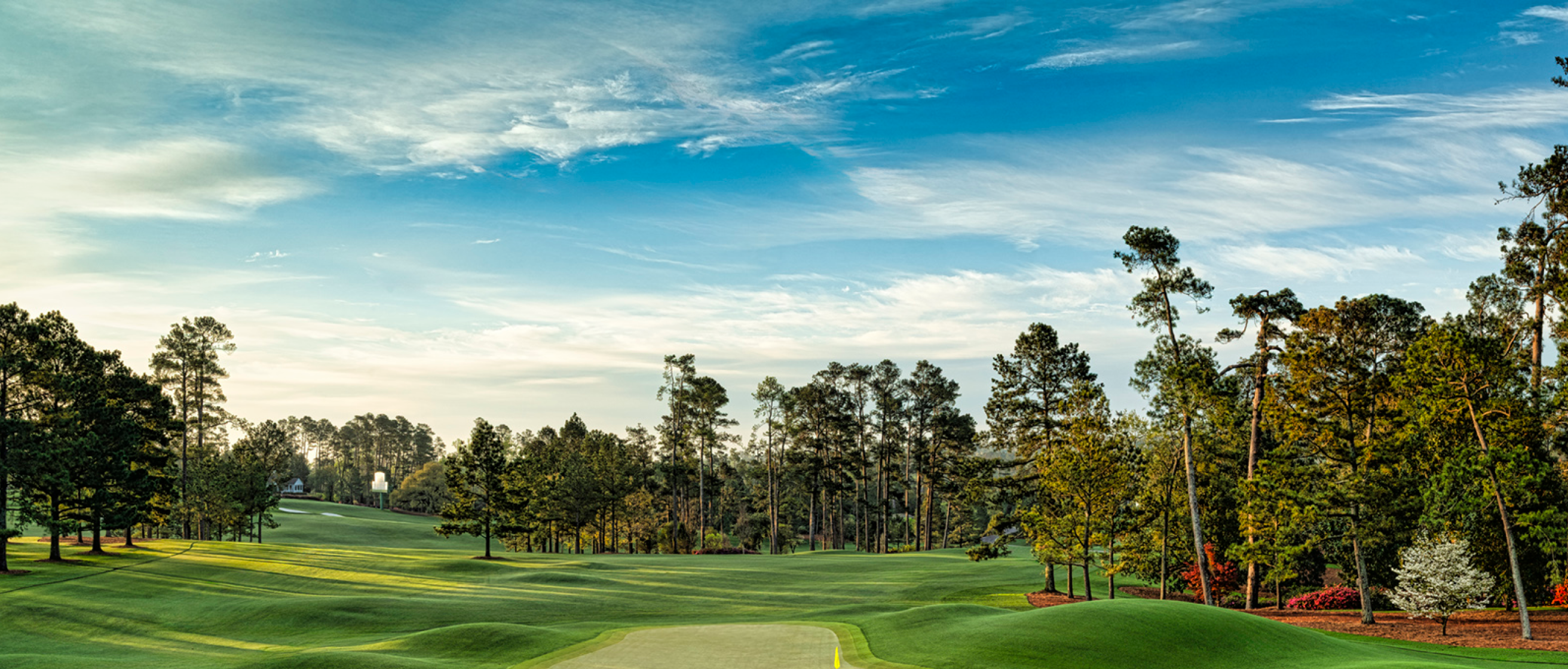Un extenso campo de golf verde bordeado por altos árboles bajo un cielo azul con nubes dispersas.