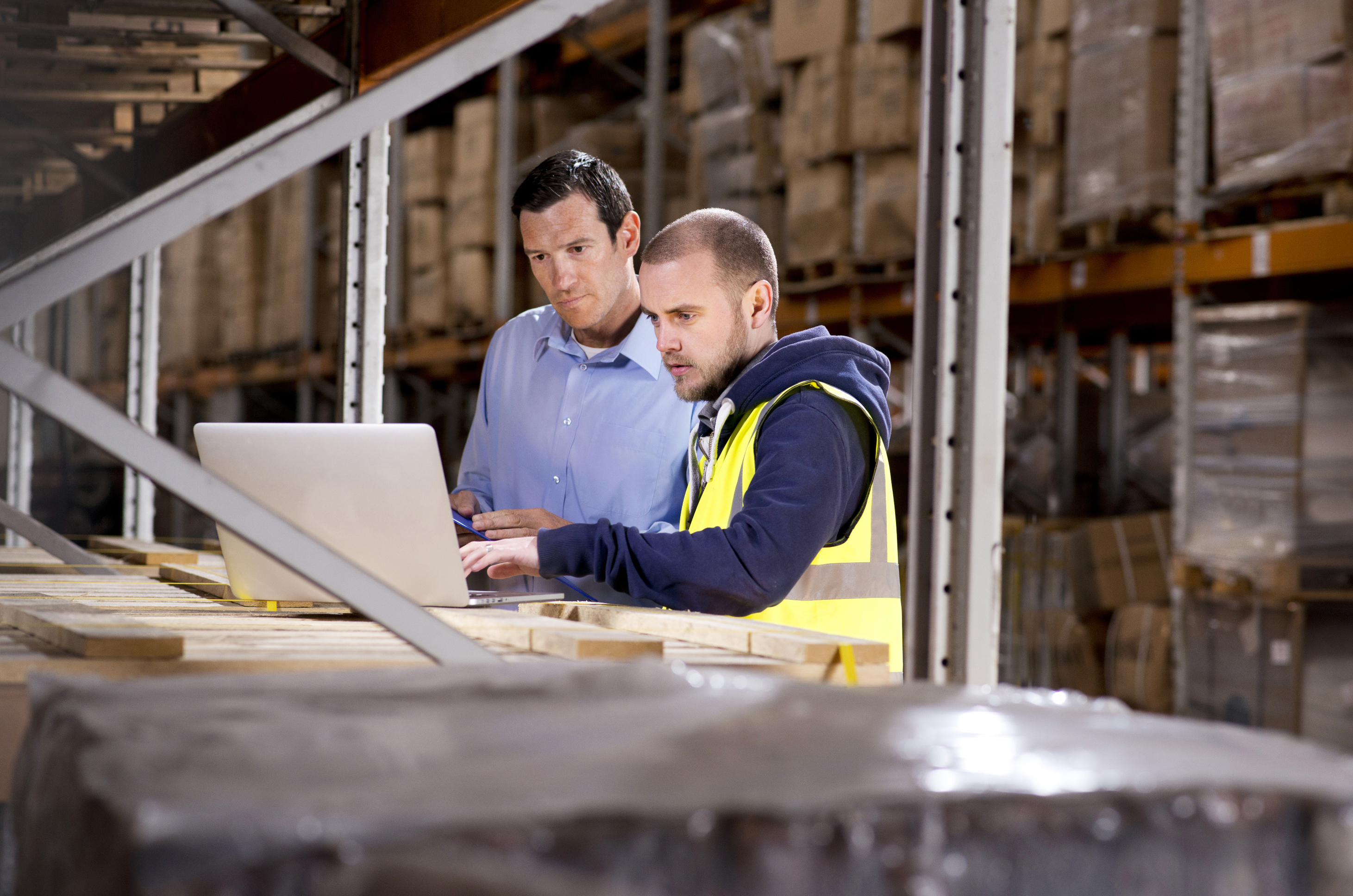 Two professionals reviewing inventory and procurement data on a laptop in a warehouse, supporting B2B commerce and purchasing workflows.